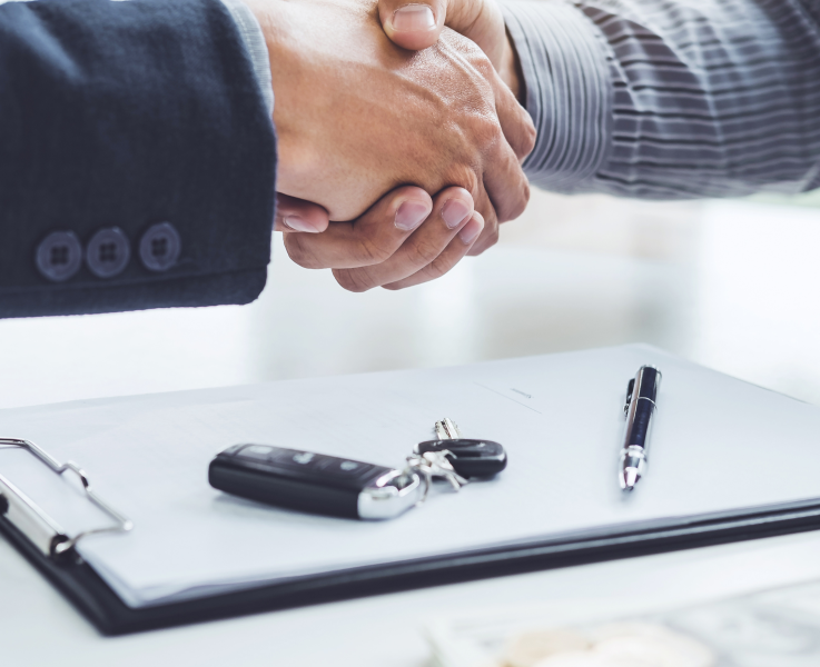 Close-up of two people in business attire shaking hands over a desk with a car key, contract on a clipboard, and pen, symbolizing closing a car purchase or financing deal.