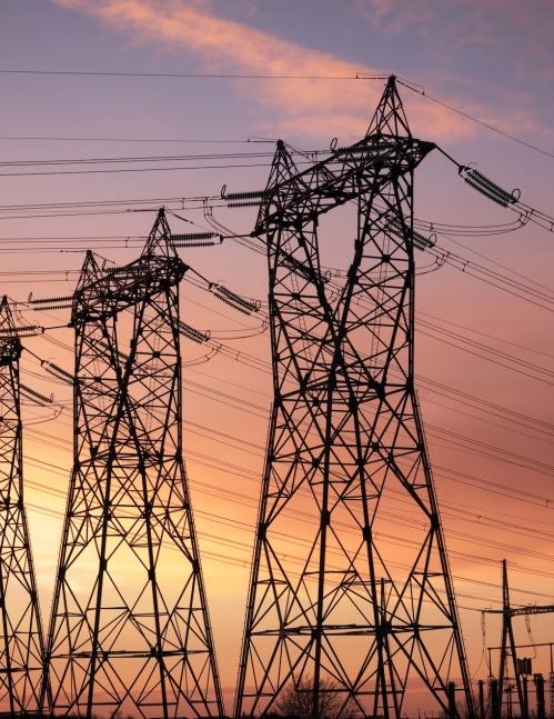 Silhouettes of a row of large steel high-voltage electricity transmission towers receding into the distance, with high-tension power lines stretching between them, set against a dramatic pink, orange, and purple sunset sky. A utility substation is visible in the lower right corner. 