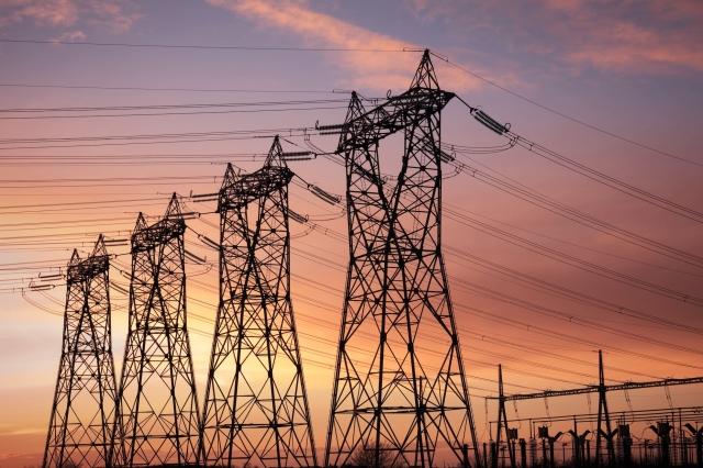 Silhouettes of a row of large steel high-voltage electricity transmission towers receding into the distance, with high-tension power lines stretching between them, set against a dramatic pink, orange, and purple sunset sky. A utility substation is visible in the lower right corner. 