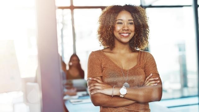 Smiling professional standing with arms crossed in a bright modern office, with coworkers blurred in the background