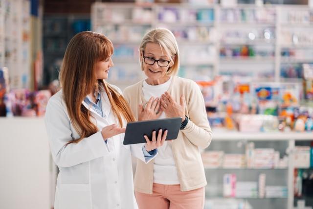 Female pharmacist in white coat showing digital tablet to senior woman customer in pharmacy, with shelves of medications visible in background