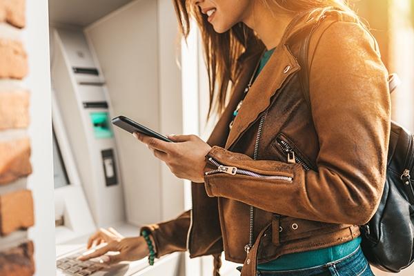 Young woman managing her finances at an ATM using a smartphone and mobile banking app