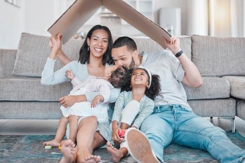 Happy family of four sitting on living room floor in front of gray couch, with parents holding cardboard roof shape over two young children, symbolizing home and family protection