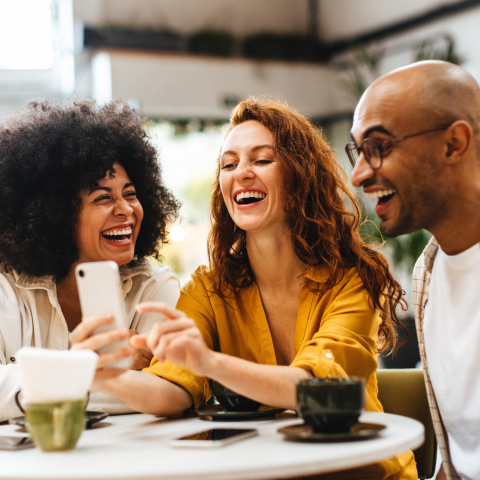 Three diverse friends laughing together at cafe table while looking at smartphone, with coffee cups on white round table in bright modern coffee shop with plants and shelving in background