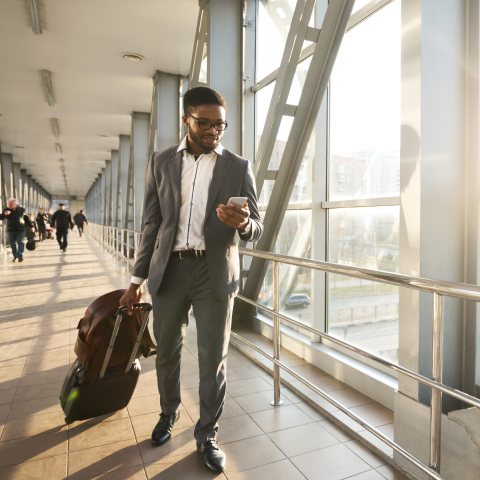 Business traveler in suit walking through bright modern airport terminal corridor with rolling luggage, checking smartphone while passing floor-to-ceiling windows with sunlight streaming in