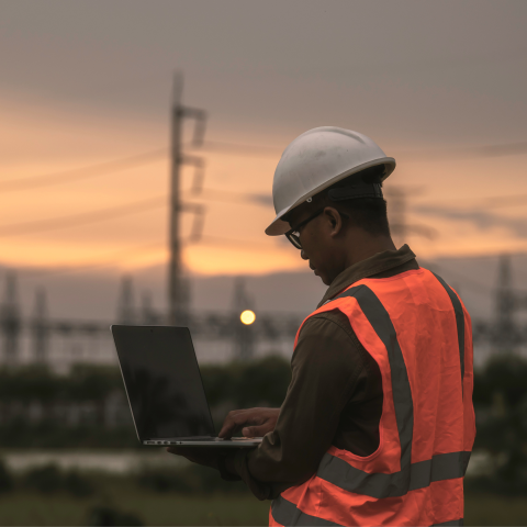 Industrial worker in white hard hat and orange high-visibility safety vest using laptop at oil refinery or chemical plant during sunset, with industrial towers and infrastructure visible in blurred background