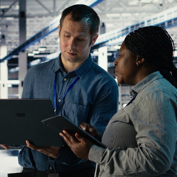 A man and a woman, both wearing lanyards, look at a laptop and tablet respectively in office.