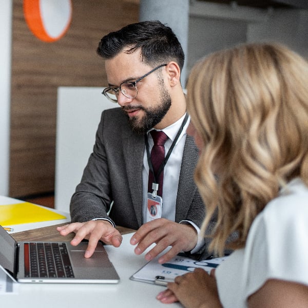 A man in a suit and glasses works on a laptop next to a woman looking at a clipboard at a desk.