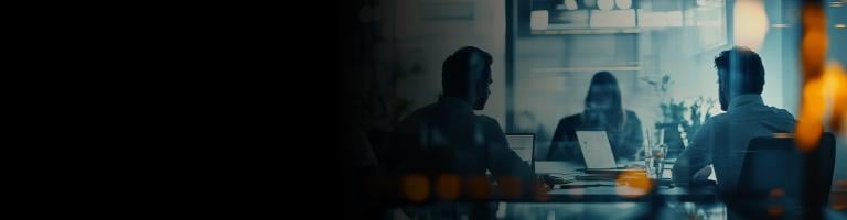 Three people working on laptops in a dimly lit meeting room, seen through a reflective glass partition.