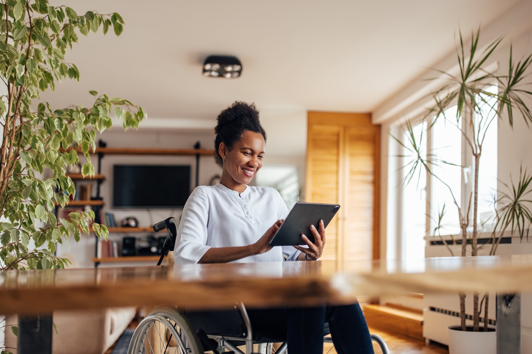 A smiling Black woman in a wheelchair uses a tablet at a wooden table in a well-lit room with plants and a window.