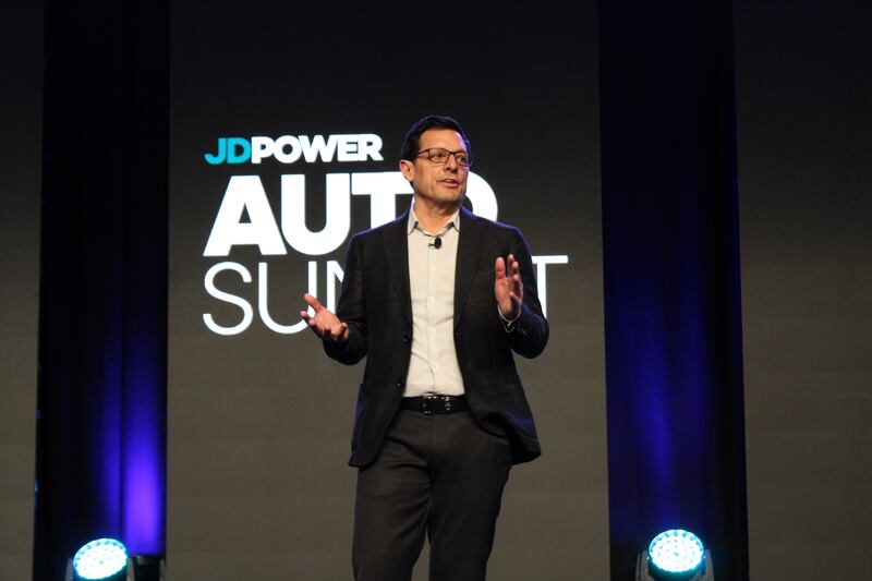 Man in a blazer and glasses speaking on stage with a 'JDPOWER AUTO SUMMIT' logo on a screen behind him, illuminated by blue lights.