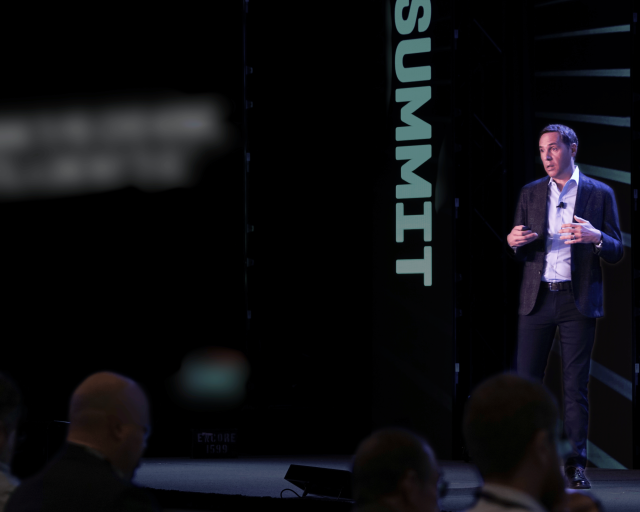 A man in a suit jacket speaks on a stage, gesturing with his hands, with the word "SUMMIT" displayed vertically on a dark screen behind him.