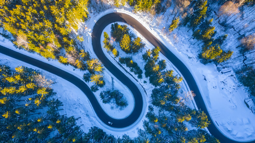 Asphalt road serpentine in snowy wintertime. Cold winter and sunny day above forest road with illuminated trees by rising sun. Aerial view from drone.|