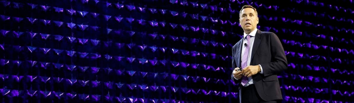 A man in a dark suit speaks on a stage with white chairs and a glowing purple geometric background.