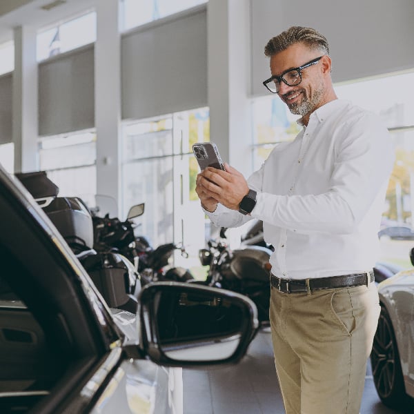 Man at dealership holding phone while looking over a vehicle.