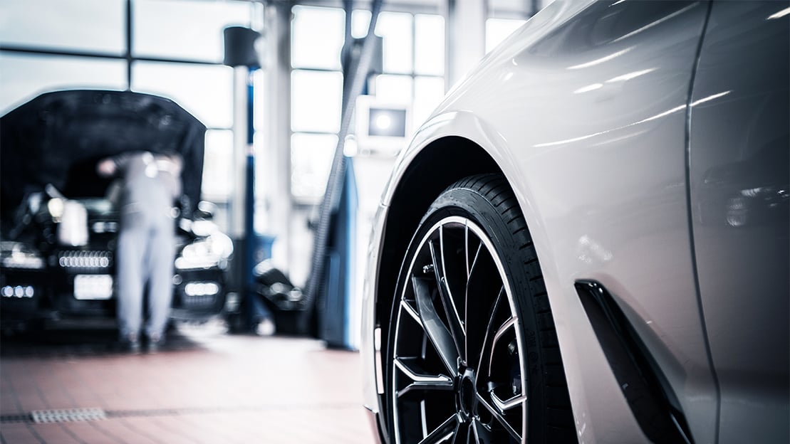 Close-up of a white car's front wheel and fender in a blurred garage background with a mechanic working on another car.